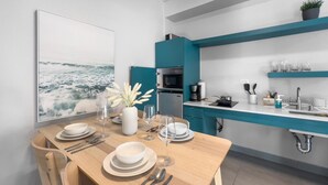 Une cuisine moderne aux meubles de couleur turquoise, une table à manger dressée avec de l’assiette blanche et des verres à vin, et un tableau représentant un paysage marin accroché au mur.