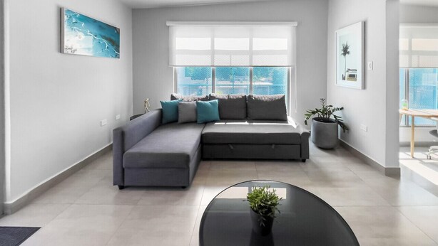 A modern living room with a sectional sofa, a round coffee table, and a large window with a view. - Aquabella #8 - Beach Bliss in Isla Verde (Carolina)