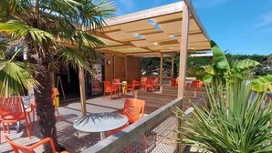 Een terras buiten met oranje stoelen en tafels, omringd door groen en een houten pergola.