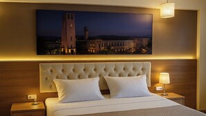 A hotel room with a large bed, a tufted headboard, two bedside tables with lamps, and a framed cityscape painting above the bed. - Lucid Otel (Izmir)