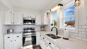A modern kitchen with white cabinets, stainless steel appliances, a large window, and pendant lighting. - Mountain View Cottage | Walk to Town | Pet (Shenandoah)
