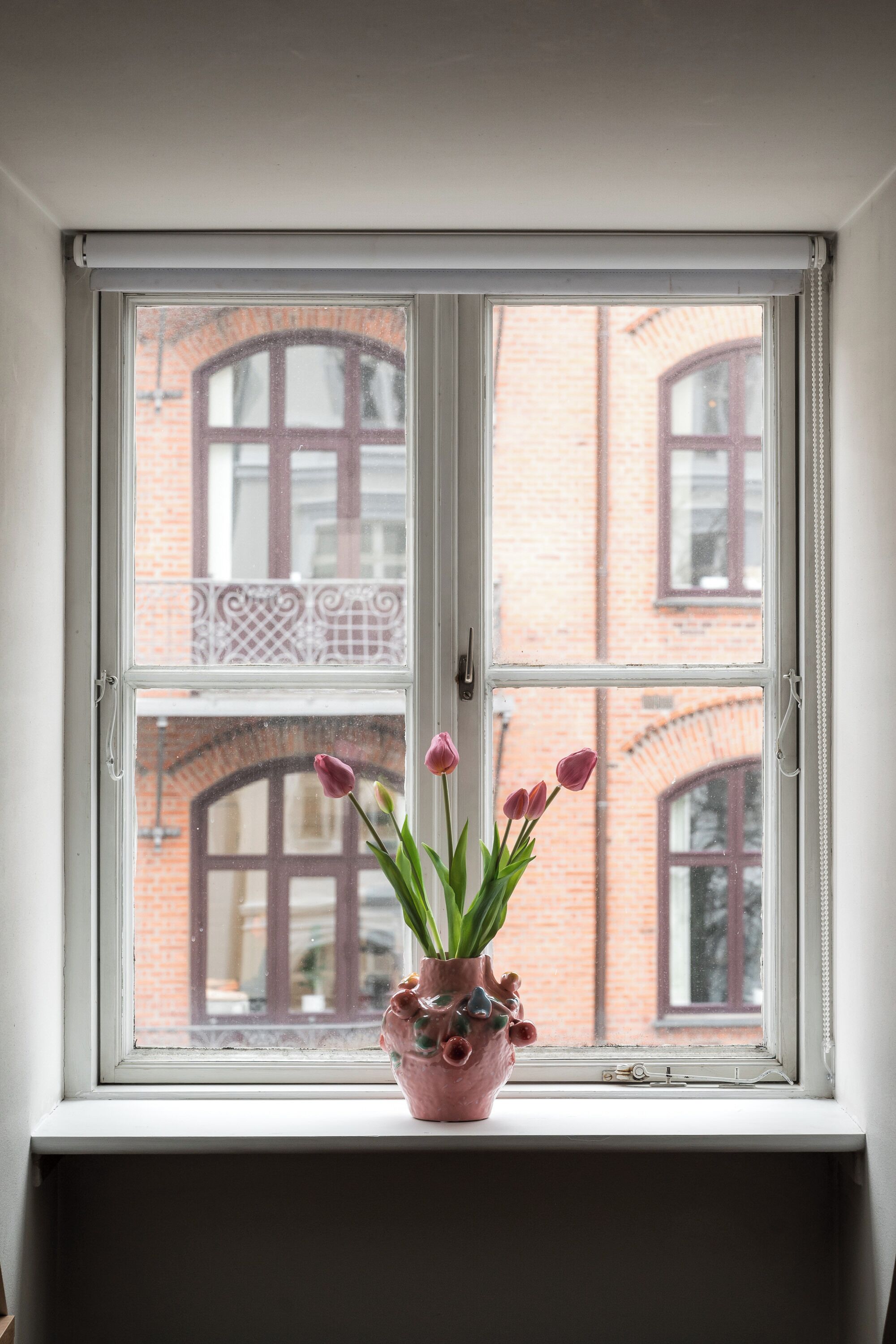 A window with a view of a brick building and a pink vase with tulips on a windowsill.
