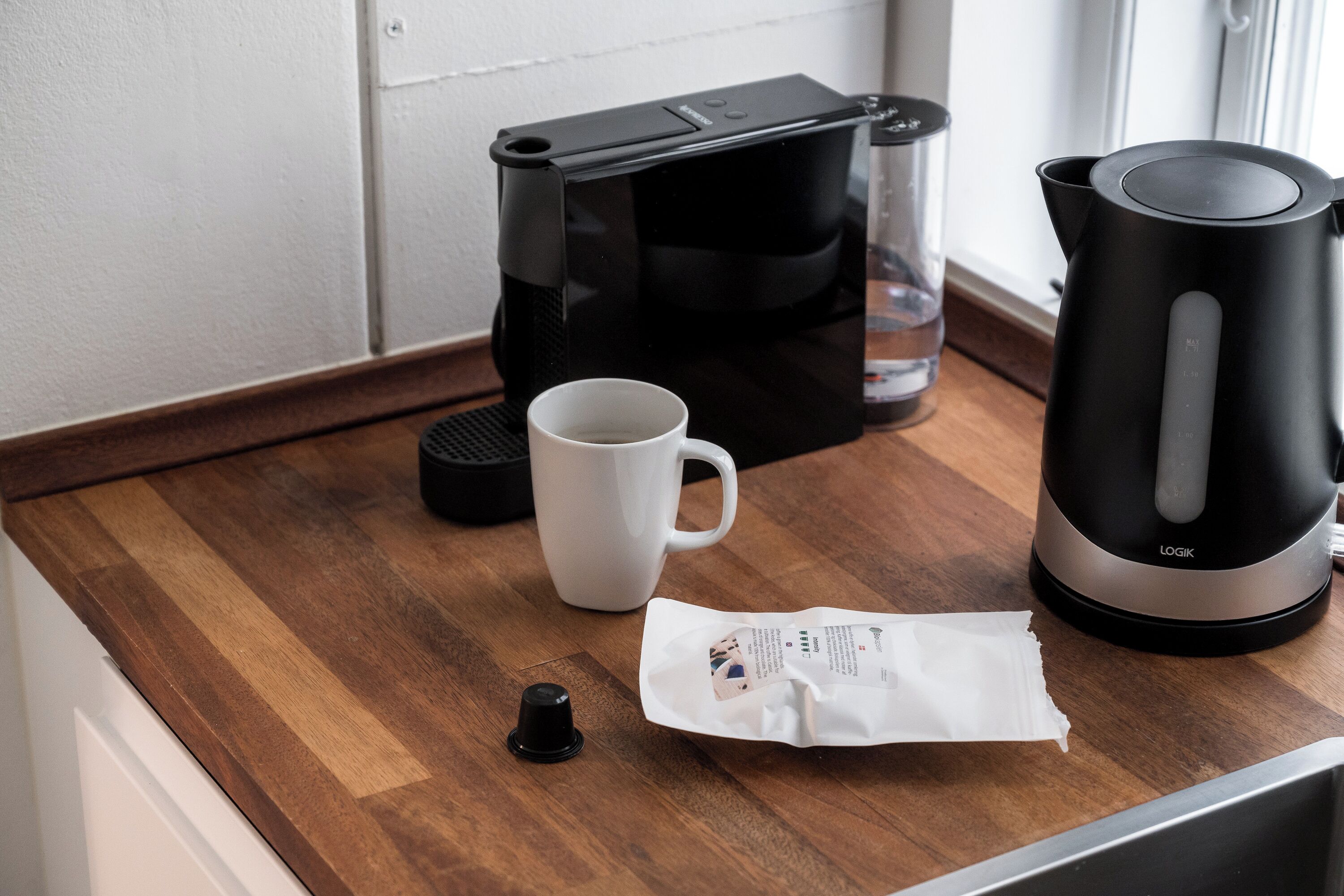 A kitchen counter with a coffee maker, a mug, a kettle, and a packet of coffee pods.