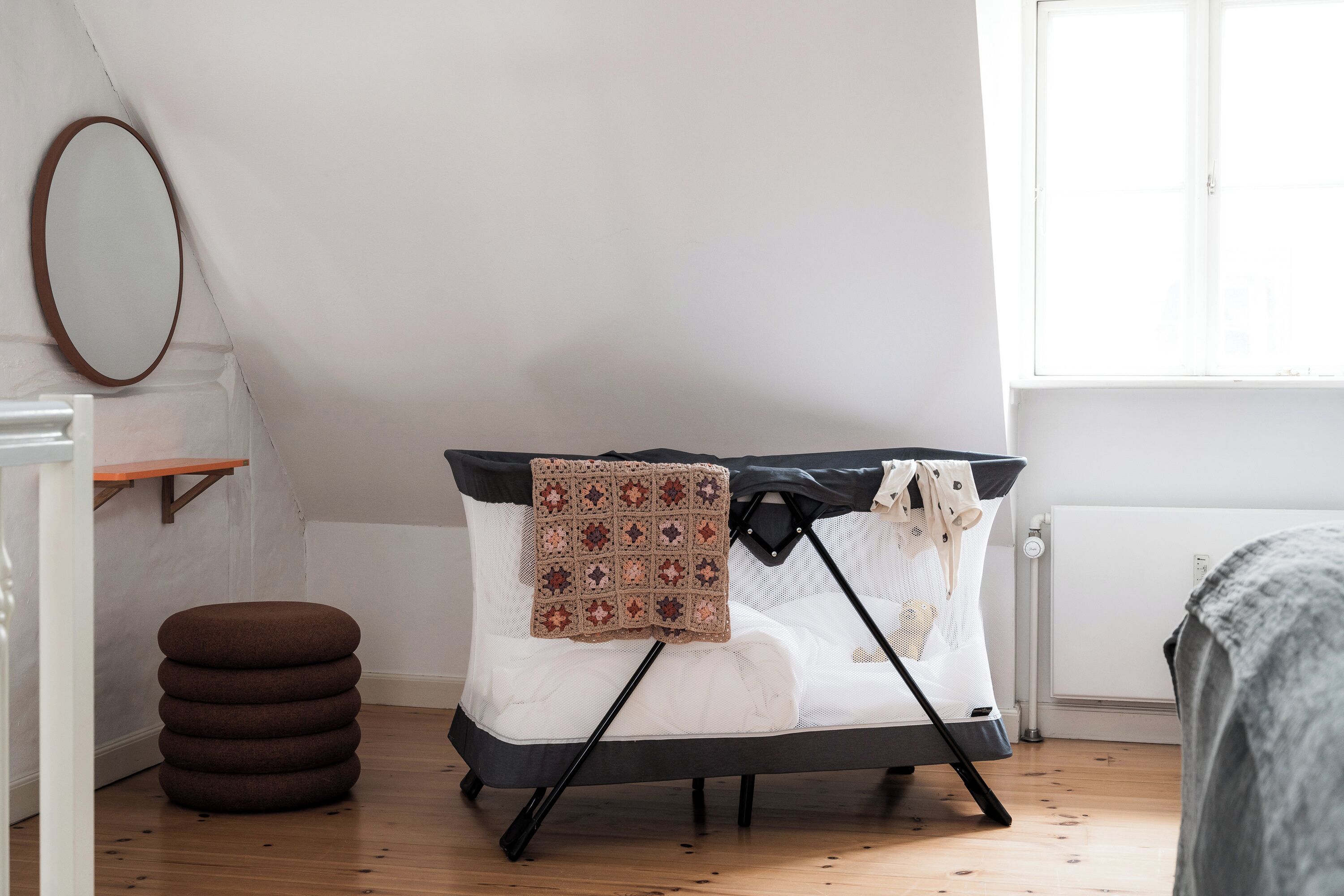 Um sofá-cama dobrável com manta estampada, um banquinho redondo e um espelho na parede.
