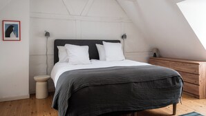 Une chambre avec un plafond en pente, un lit avec une literie blanche et gris foncé, une commode en bois et un petit tabouret.