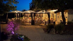 A covered outdoor seating area with tables and chairs, surrounded by trees and a building with large windows. - Holiday Home in France with Private Terrace (Mauroux)
