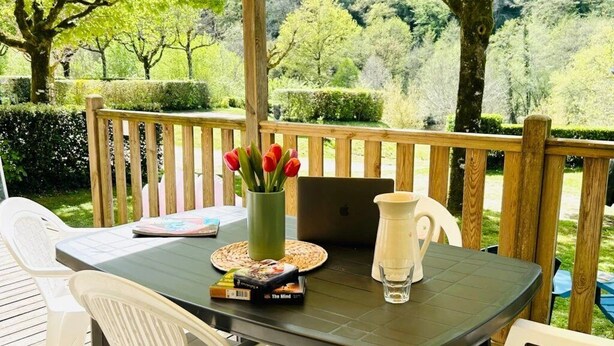 Une terrasse en bois avec une table préparée pour deux personnes, sur laquelle on trouve un ordinateur portable, un livre, une assiette de nourriture et une carafe.