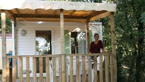 A wooden deck with a covered porch, a white building with green shutters, and a person standing on the porch. - ZEcamping 3 étoiles animé piscine chauffée dog friendly locatif BBQ (Mondragon)