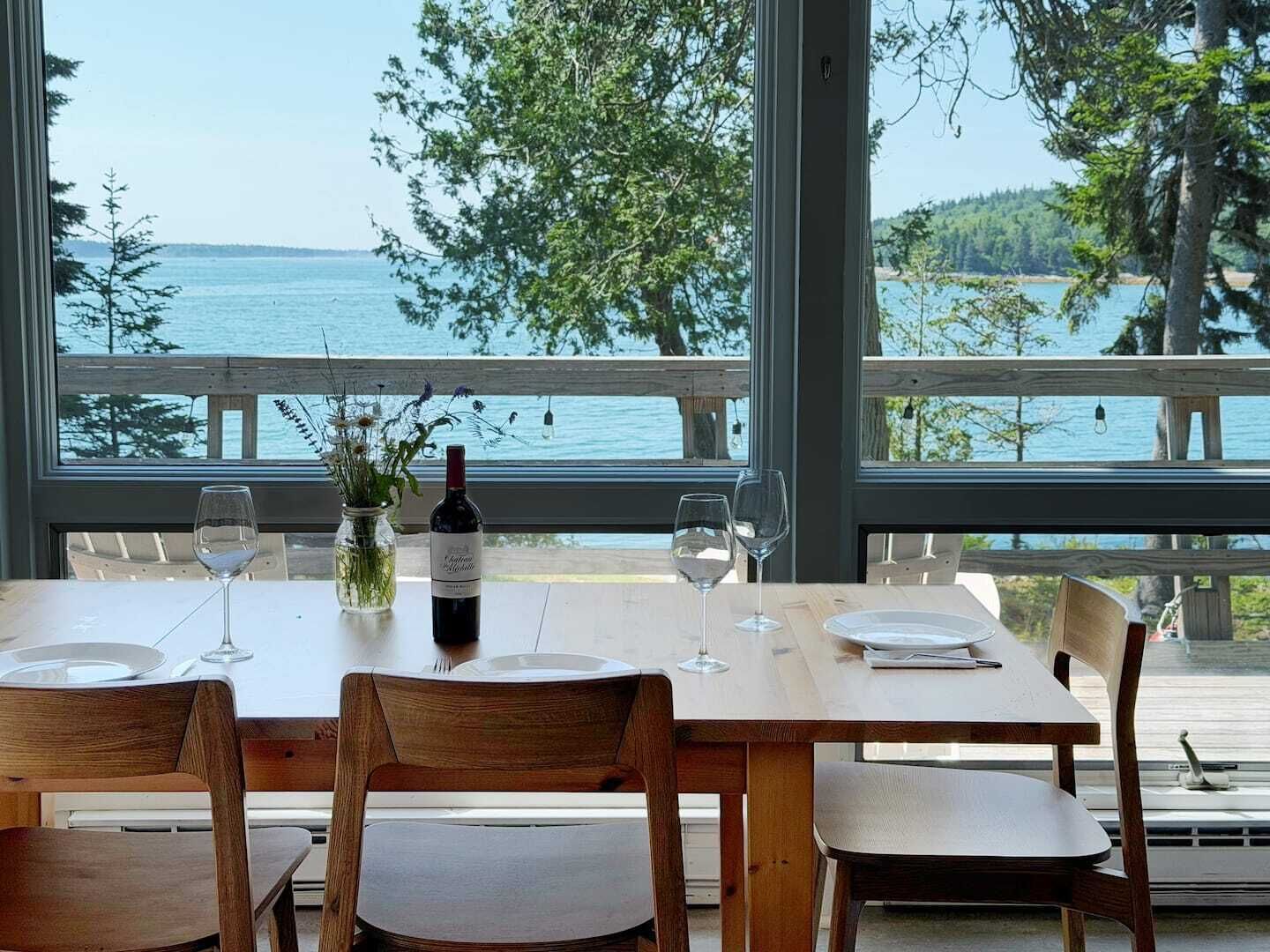 A dining table with chairs, a bottle of wine, and glasses, set against a backdrop of a lake and trees.