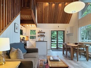 A spacious living area with a wooden staircase, a dining table with chairs, a kitchen with white cabinets, and large windows offering a view of the outdoors. - Stunning Views - Downeast Chalet with Fireplace (Steuben)