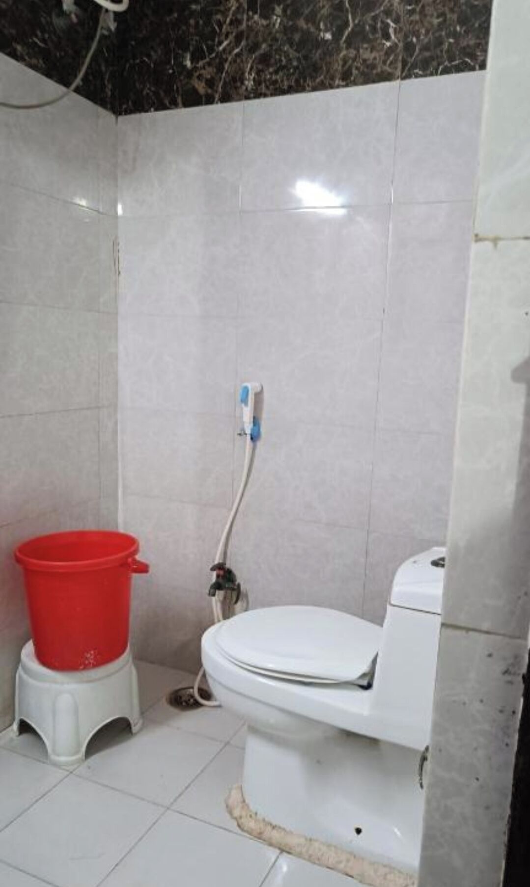 A small bathroom with a toilet, a red bucket, and a shower hose.