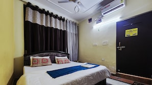A neatly made bed with a white and blue bedspread, a dark headboard, and decorative pillows. There is a ceiling fan, an air conditioning unit, and a door with a sign on it. - Divy Dhaam Near Prem Mandir (Vrindavan)