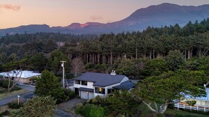 Une maison à deux étages, avec un toit sombre, entourée d’une forêt dense et avec des montagnes en arrière-plan.