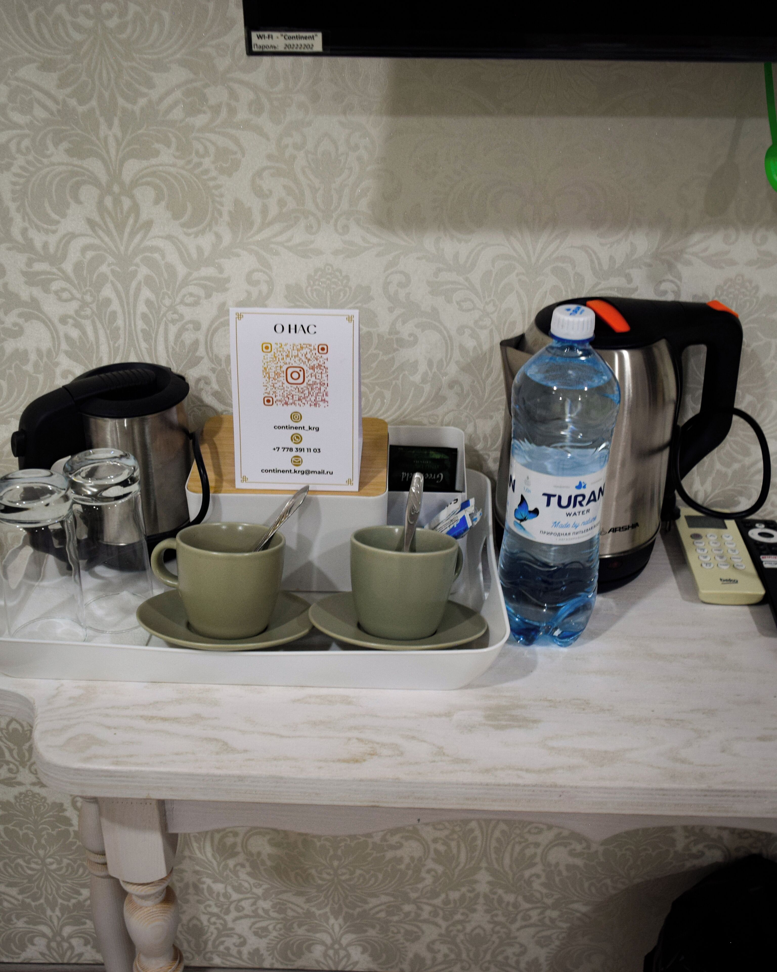 Een keukenblad met een koffiezetapparaat, twee kopjes, een waterfles, een waterkoker, een afstandsbediening en een bordje met een QR-code.