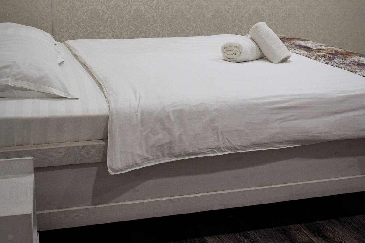 Una cama bien hecha con sábanas blancas y dos toallas enrolladas, suelo de madera y una pared con un estampado discreto.
