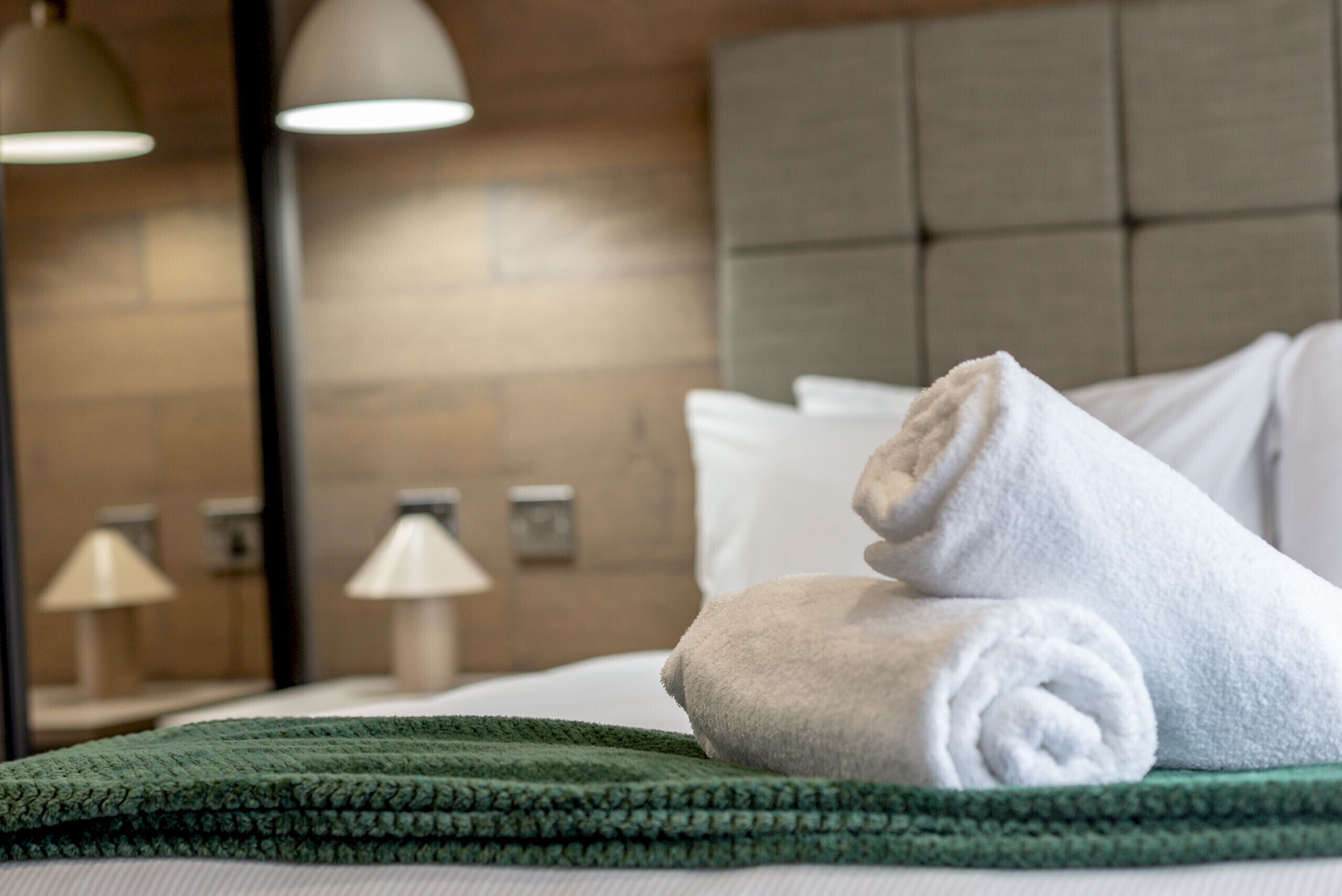 Un lit bien fait, avec des serviettes blanches pliées en rouleaux, un plaid vert et une tête de lit à motifs.