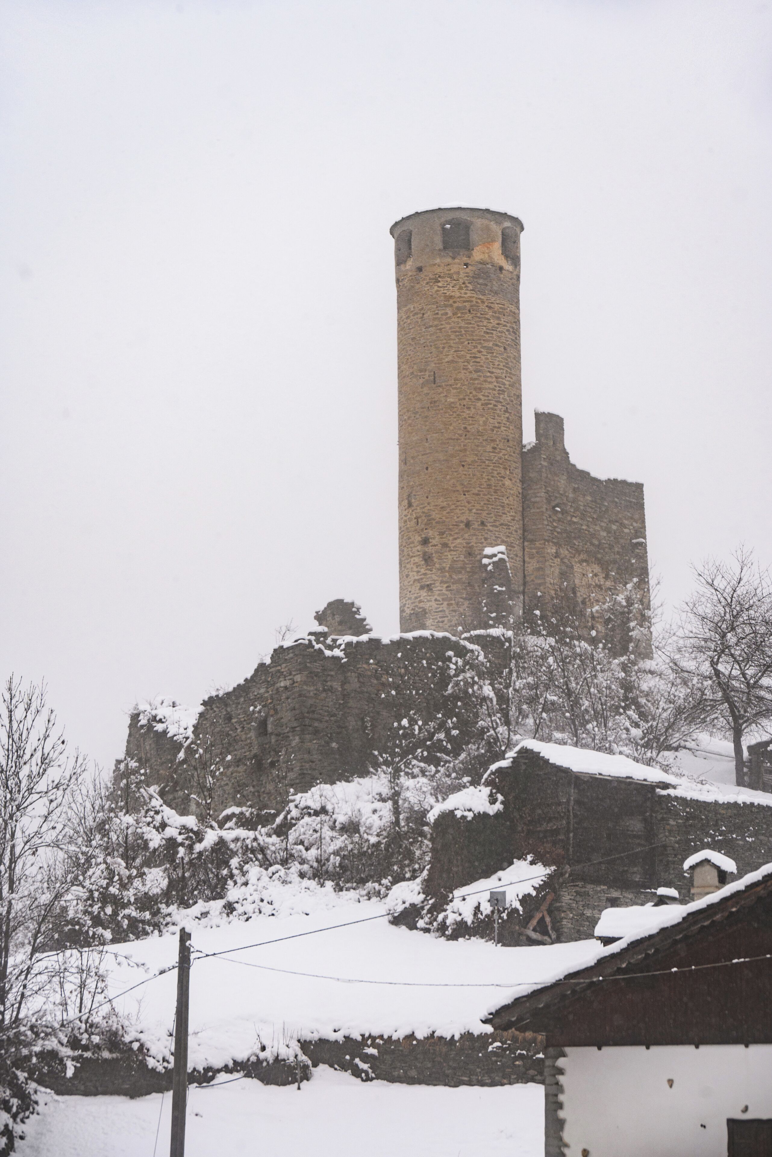 Una torre de piedra histórica de aspecto desgastado, rodeada de árboles y edificios cubiertos de nieve.