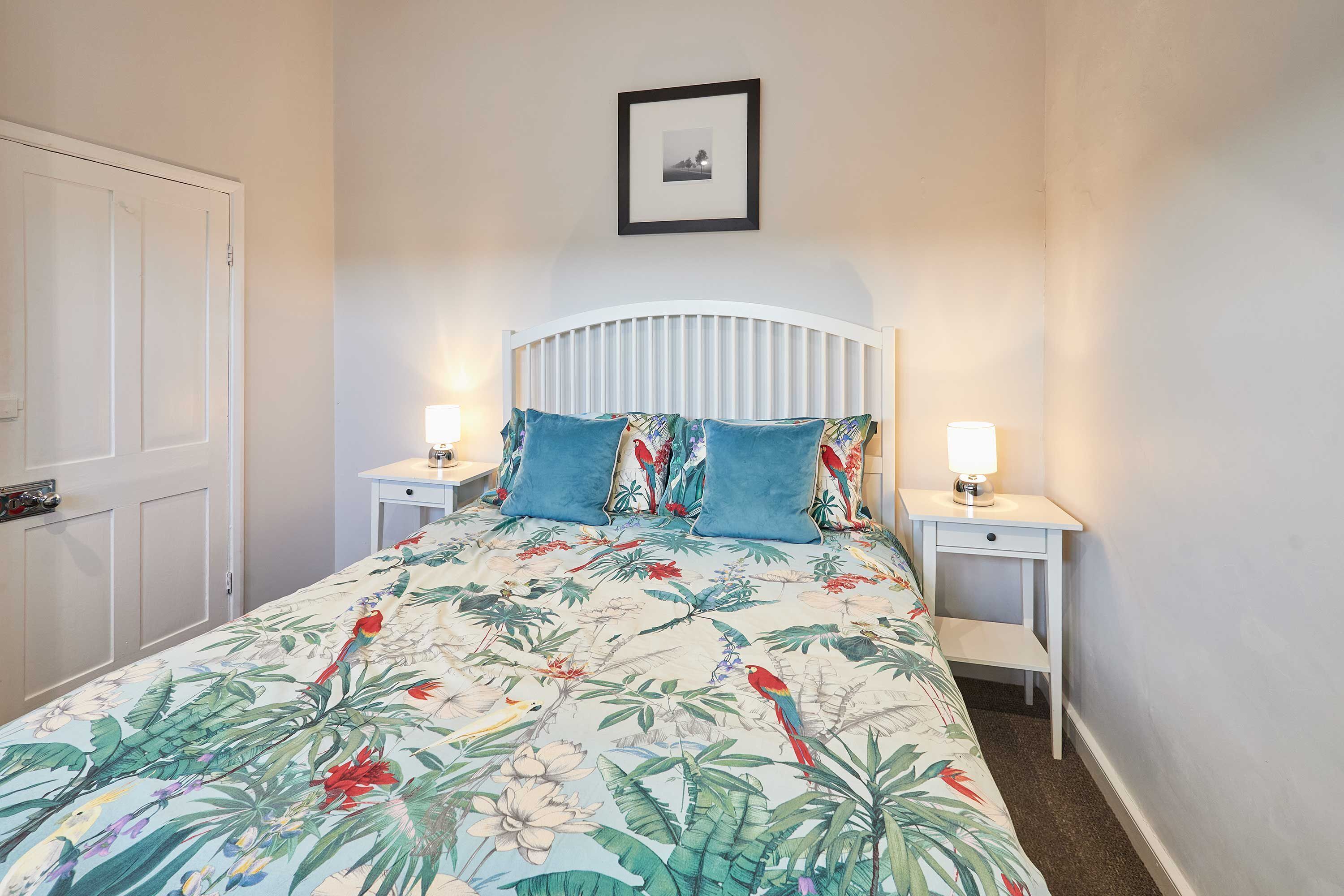 A bedroom with a bed, two bedside tables, a nightstand, a framed picture on the wall, and a door.