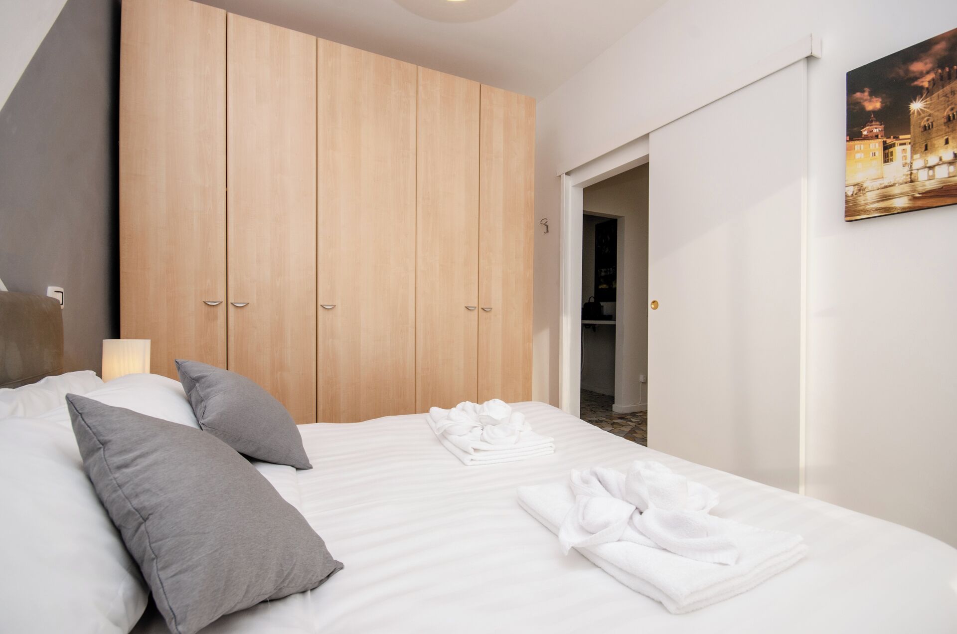 A hotel room with a large bed, grey pillows, a wooden wardrobe, and a painting of a cityscape.