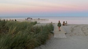 A beach scene with a wooden boardwalk leading to the water, grassy dunes, and a sunset sky. - Charming home in Marielyst - Bøtø (Væggerløse)