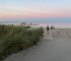 A beach scene with a wooden boardwalk leading to the water, grassy dunes, and a sunset sky. - Charming home in Marielyst - Bøtø (Væggerløse)
