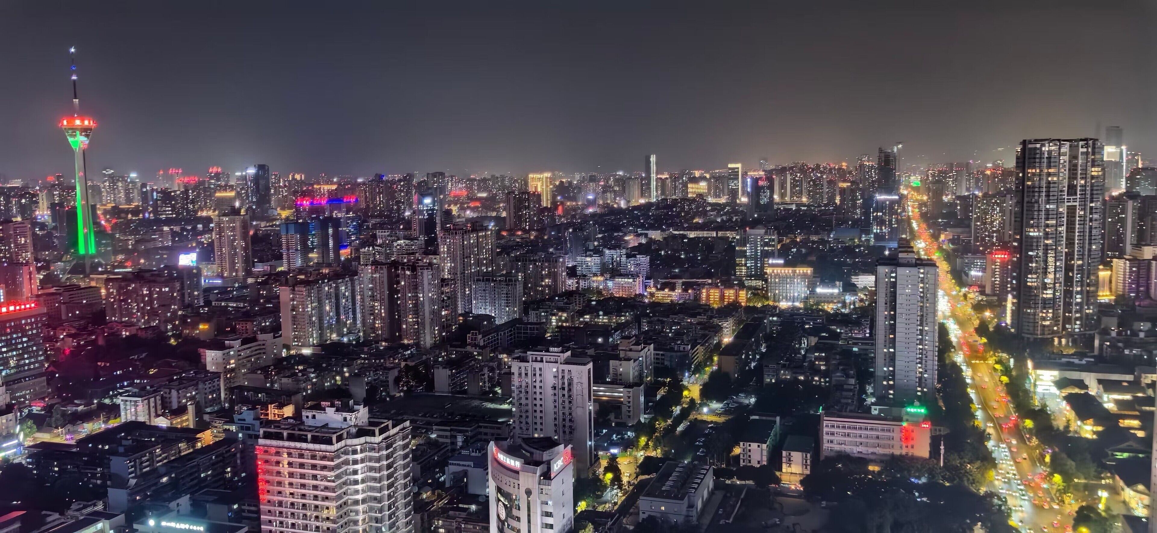 A nighttime cityscape with a prominent tower lit in green and red, surrounded by densely packed high-rise buildings and busy streets.