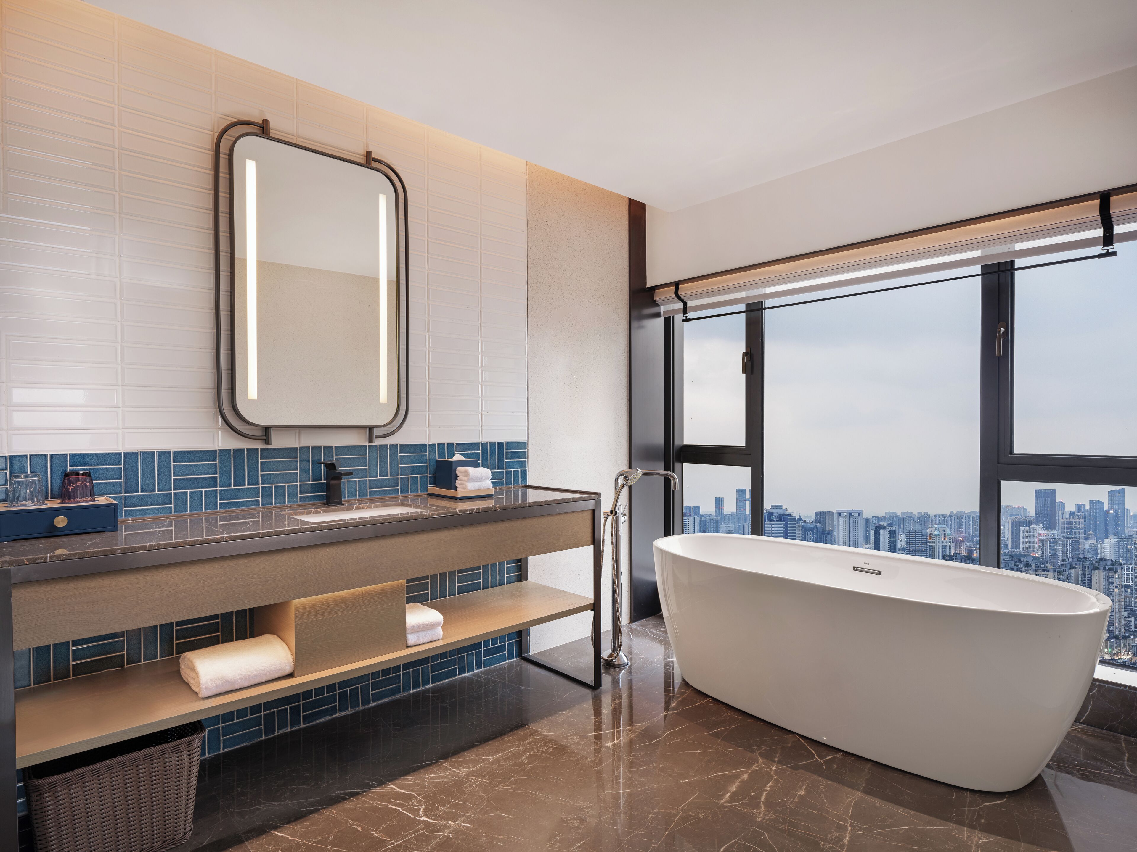 A modern bathroom with a large bathtub, a vanity with a mirror, and a city view through large windows.