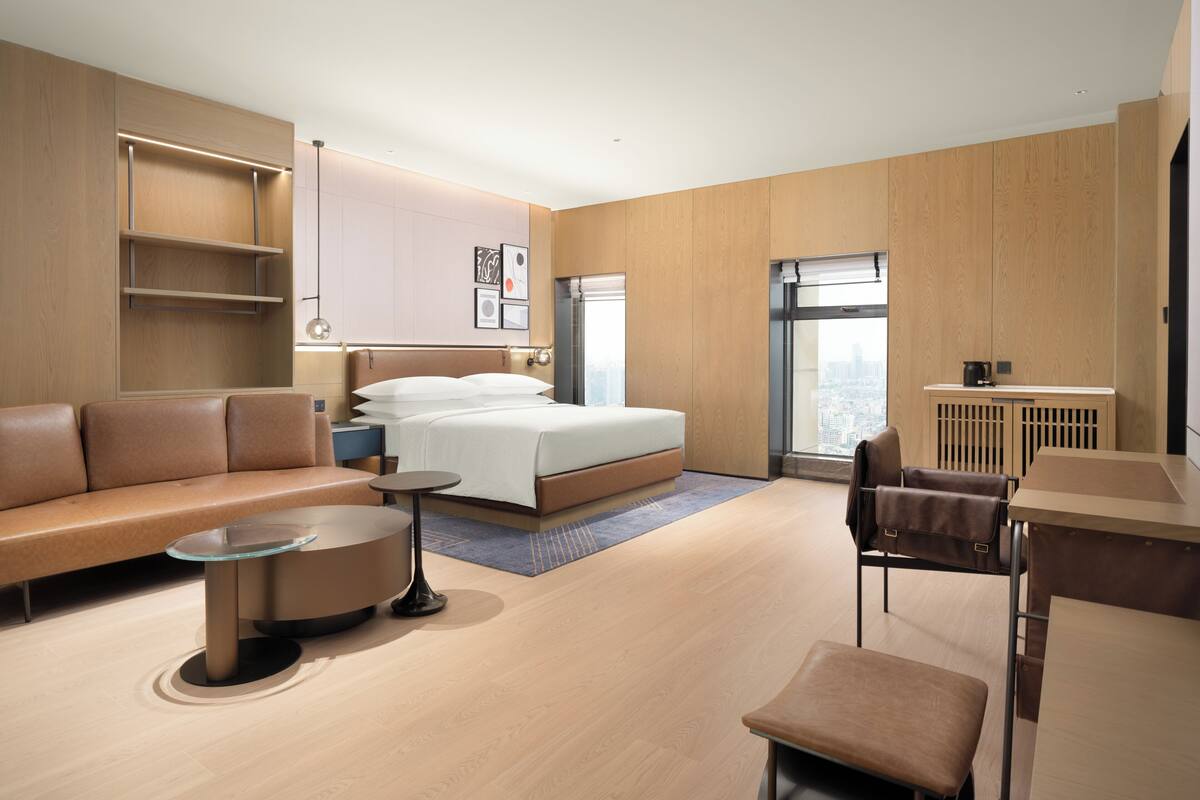 A modern hotel room with a bed, a sofa, a desk, and a view of the city.