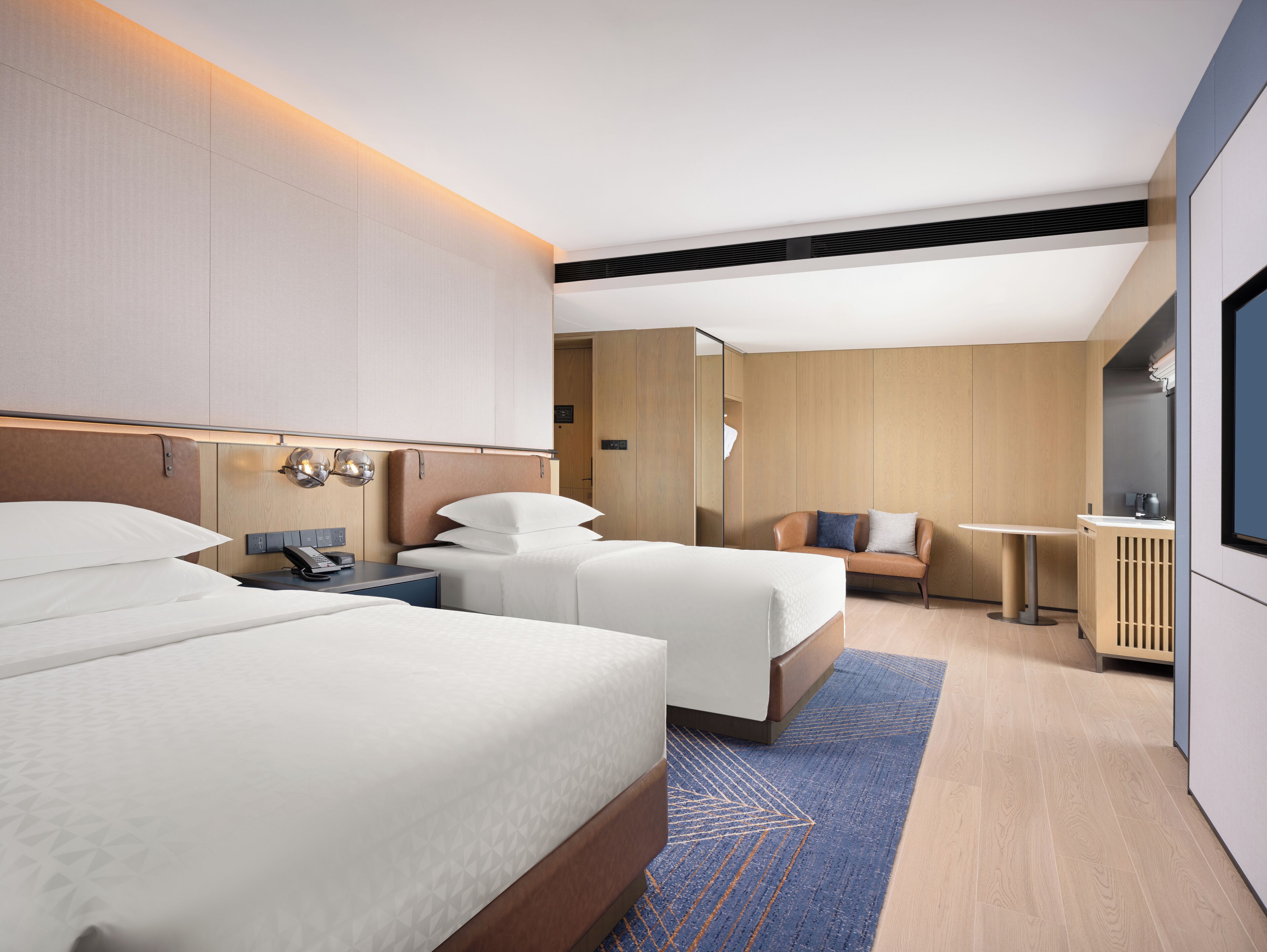 A modern hotel room with two beds, a small seating area, and a TV.