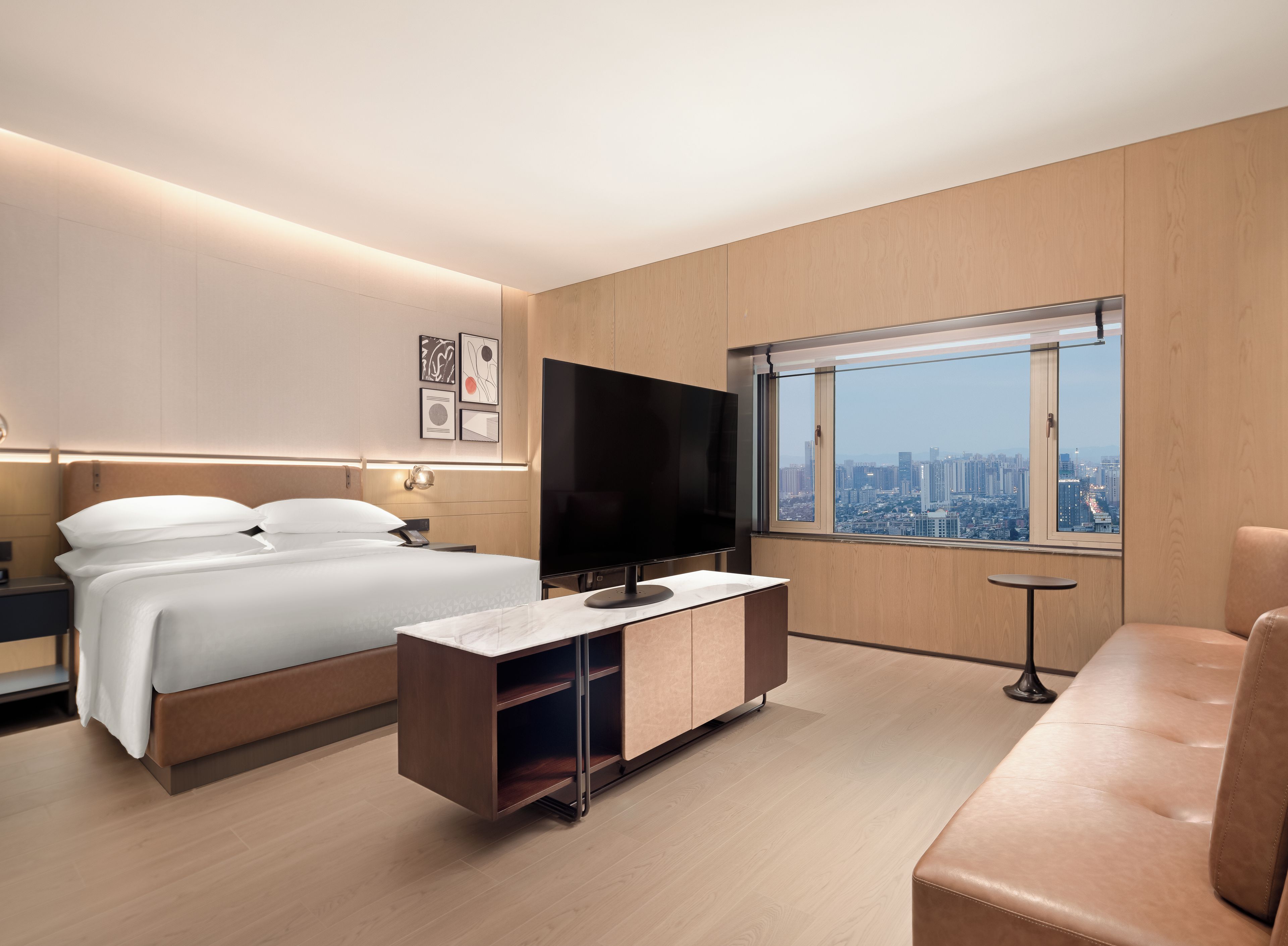 A modern hotel room with a bed, a TV, a seating area, and a city view.