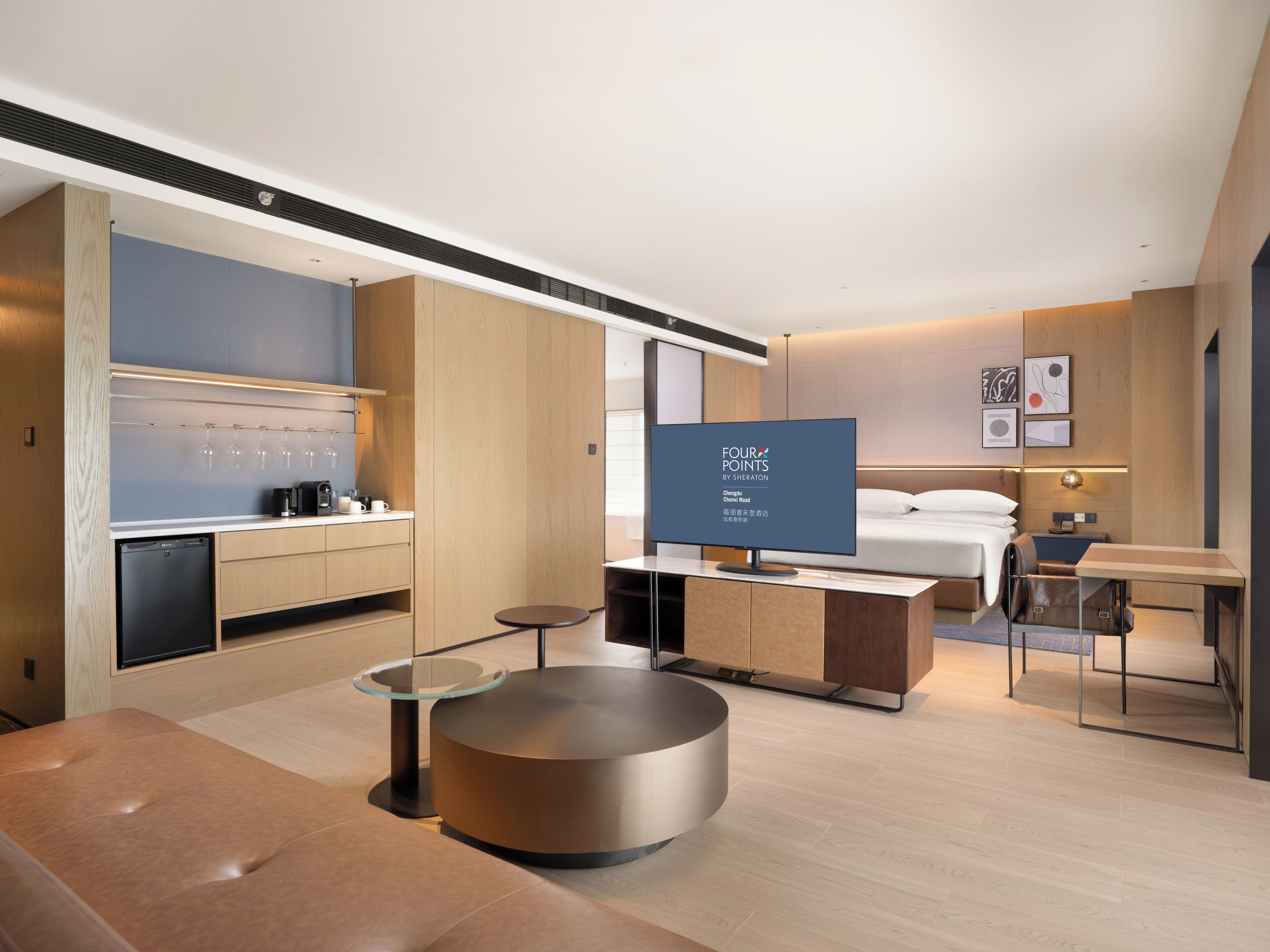 A modern hotel room with a kitchenette, a round seating area, a bed, and a television displaying "FOUR POINTS BY SHERATON".