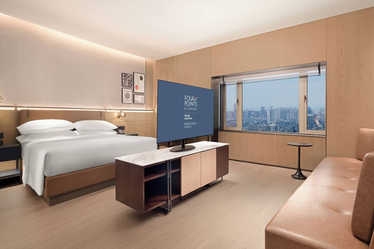 A modern hotel room with a bed, a TV, a desk, and a view of the city.