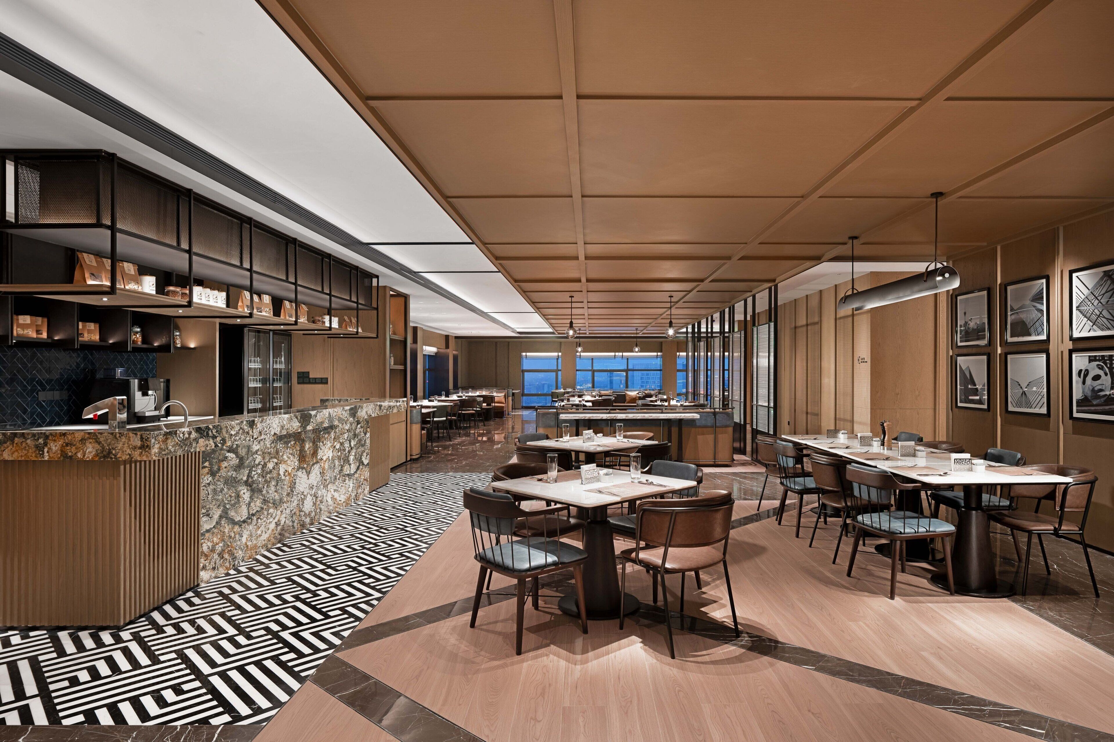 A modern dining area with a long counter, multiple tables, and chairs. There are shelves with items on the left, and a patterned floor.
