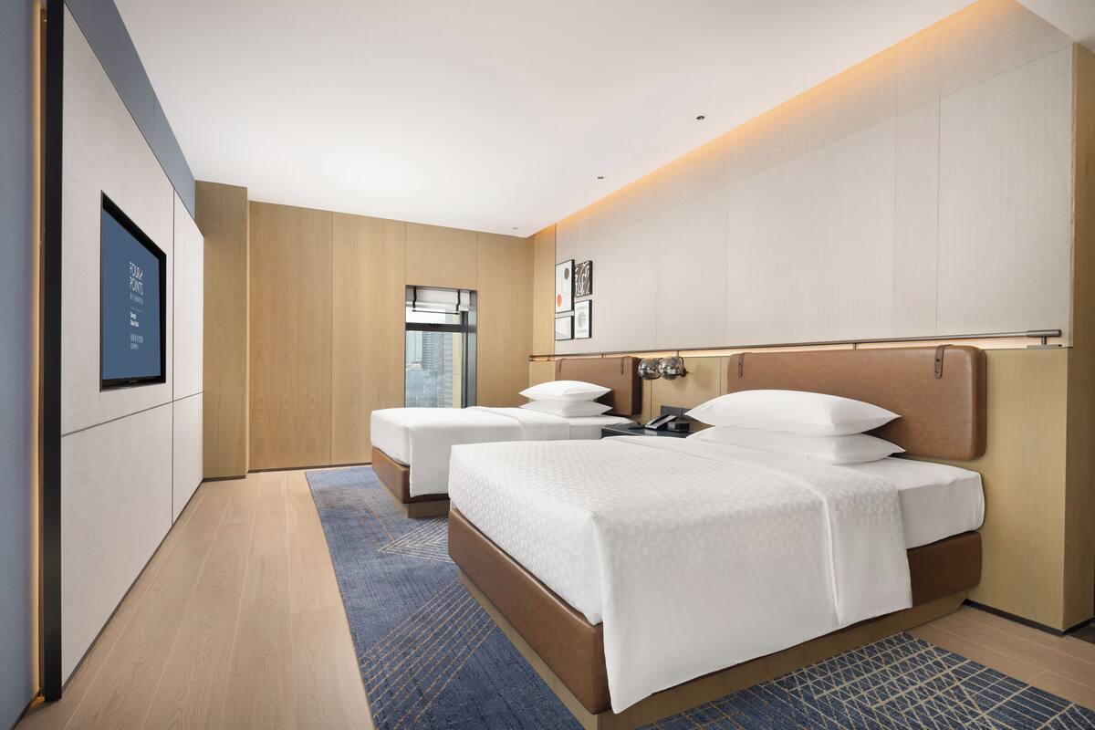 A modern hotel room with two beds, a large window, and a view of the city.