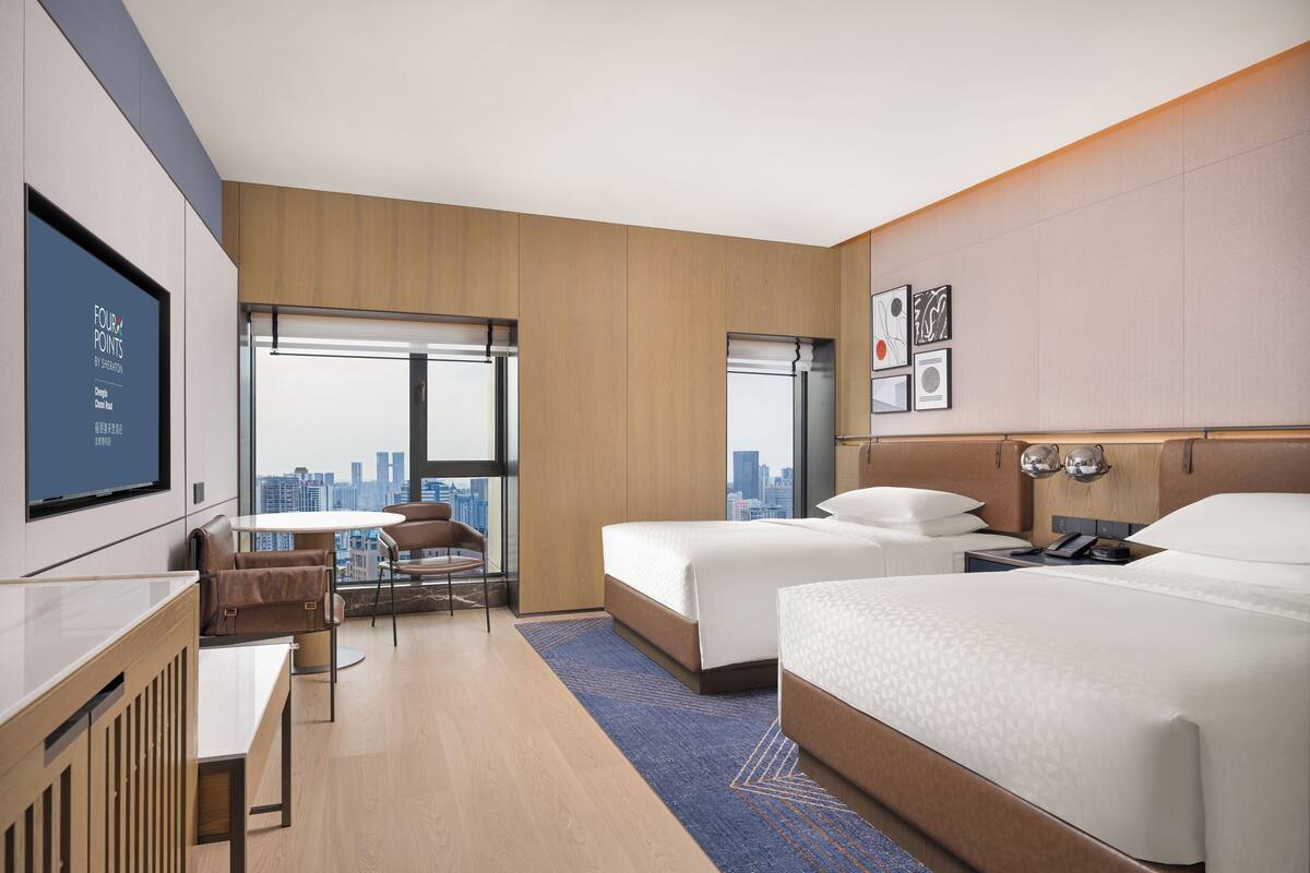 A hotel room with two beds, a desk, a chair, a TV, and a city view.