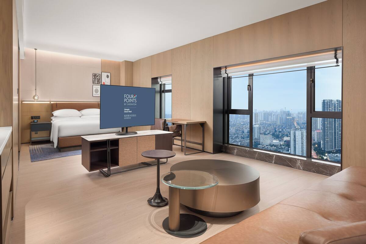 A modern hotel room with a bed, a desk, a glass table, a sofa, and a large window with a city view.