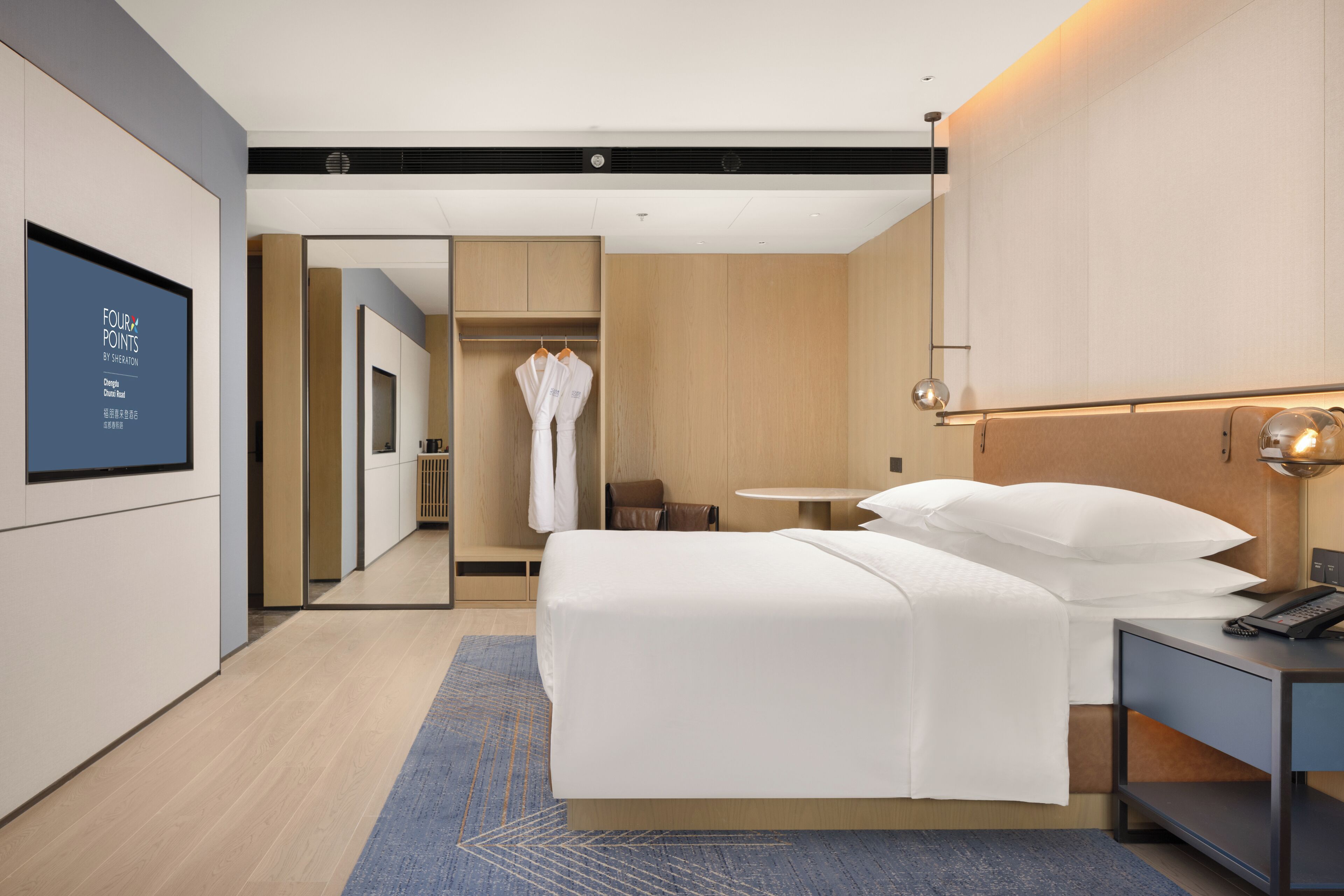A modern hotel room with a large bed, bedside table, and a mirror above it. There is a small table and a chair in the corner, and a robe hanging on a rack.