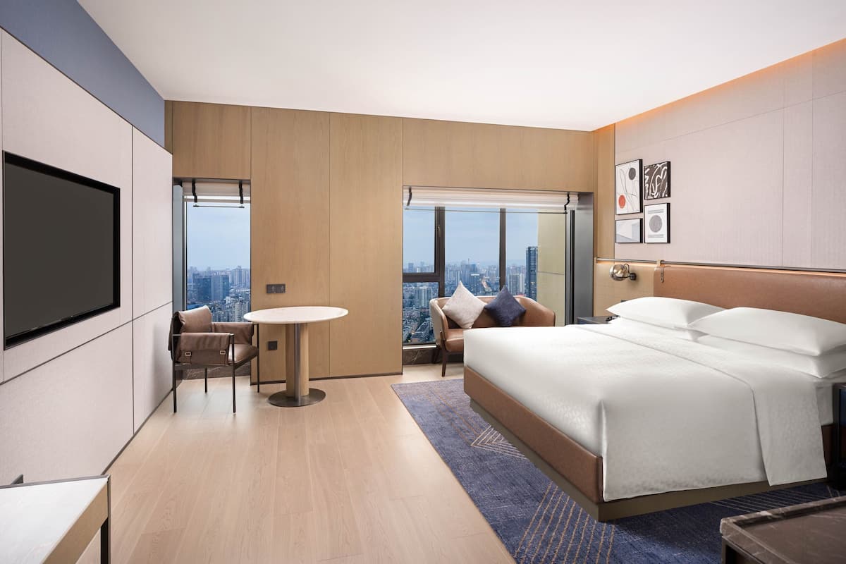 A modern hotel room with a large bed, a desk, a chair, a round table, and a city view through a large window.