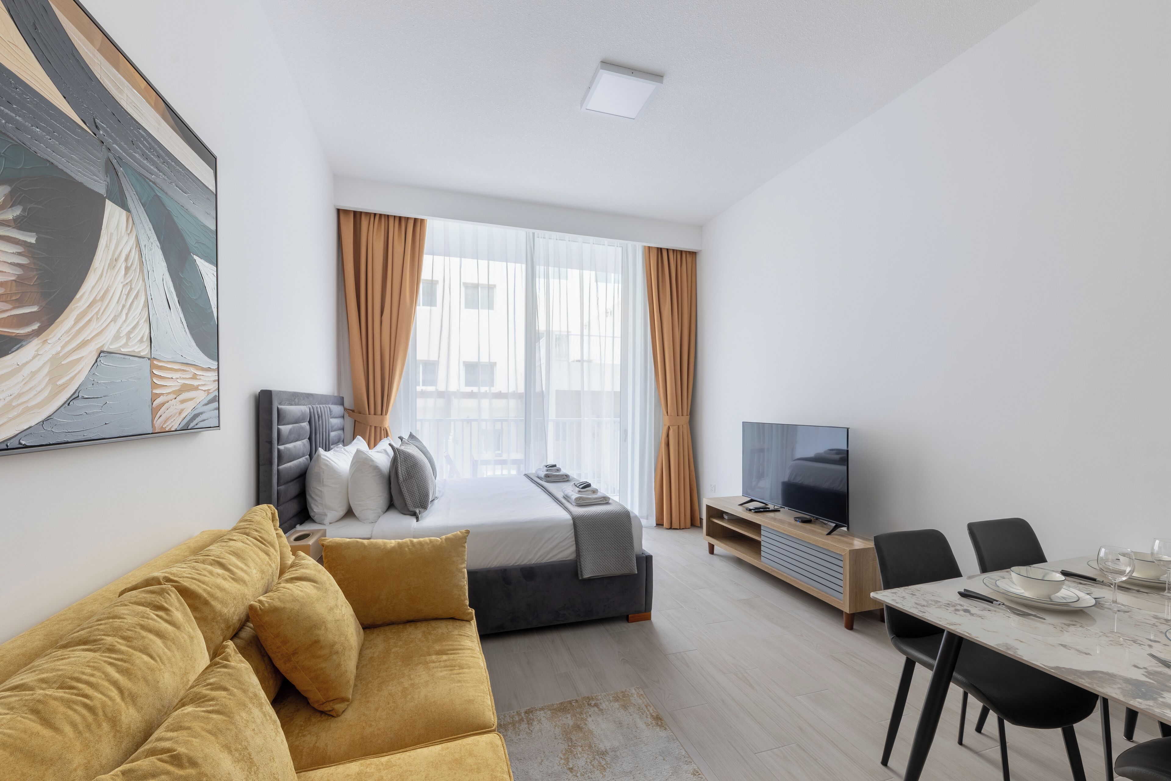 A modern hotel room with a bed, sofa, dining table, and TV.
