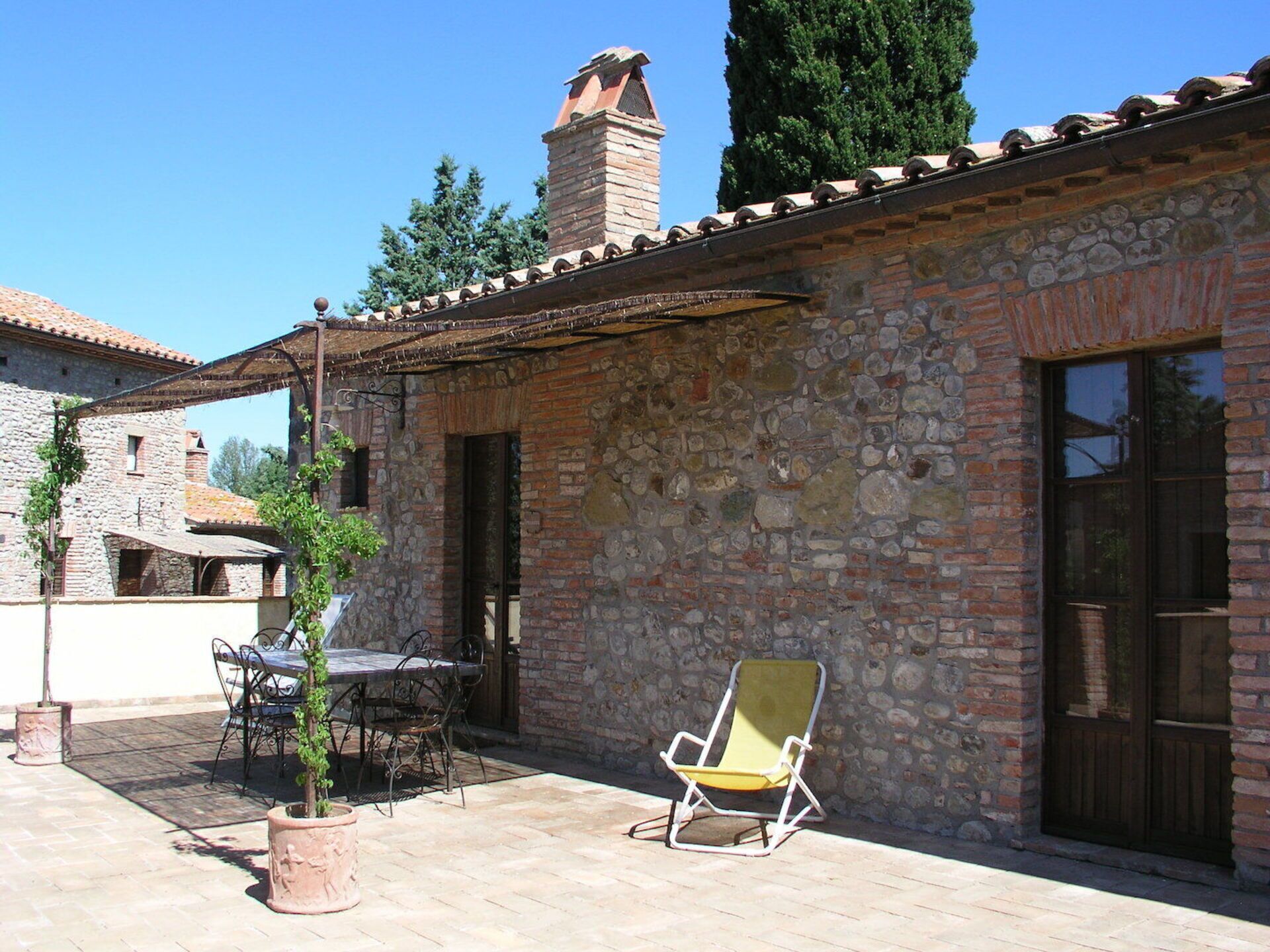 A stone building with a tiled roof, a chimney, and a patio area with a table, chairs, and a sun lounger.