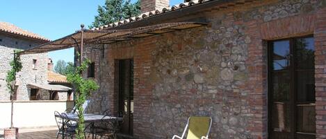 A stone building with a tiled roof, a chimney, and a patio area with a table, chairs, and a sun lounger.