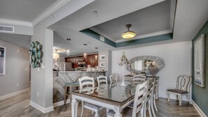 A dining area with a glass-top table surrounded by white chairs, a chandelier, and a decorative mirror on the wall.