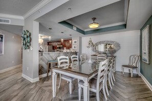 Un comedor con una mesa de vidrio rodeada de sillas blancas, una araña y un espejo decorativo en la pared.