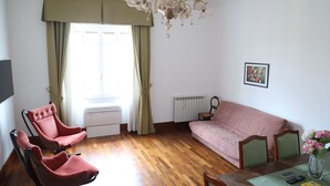 A room with a wooden floor, a sofa, two armchairs, a dining table with chairs, a chandelier, a radiator, and a window with curtains. - DOMUS HORTENSIA AT THE VATICAN (Roma)