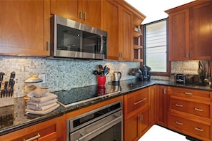 A kitchen with wooden cabinets, a stainless steel microwave, a tiled backsplash, and a window with blinds. - Wailea Elua 1304 - Spectacular Oceanfront Condo with Panoramic Views (Wailea, Maui)