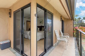 A balcony with a sliding glass door leading to a bedroom. There are two white chairs on the balcony, a small suitcase, and a trash bin.