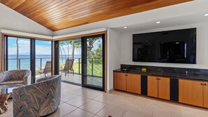 A modern kitchen with a large TV, wooden cabinets, and a view of the ocean through large sliding doors. - Wailea Elua 1304 - Spectacular Oceanfront Condo with Panoramic Views (Wailea, Maui)