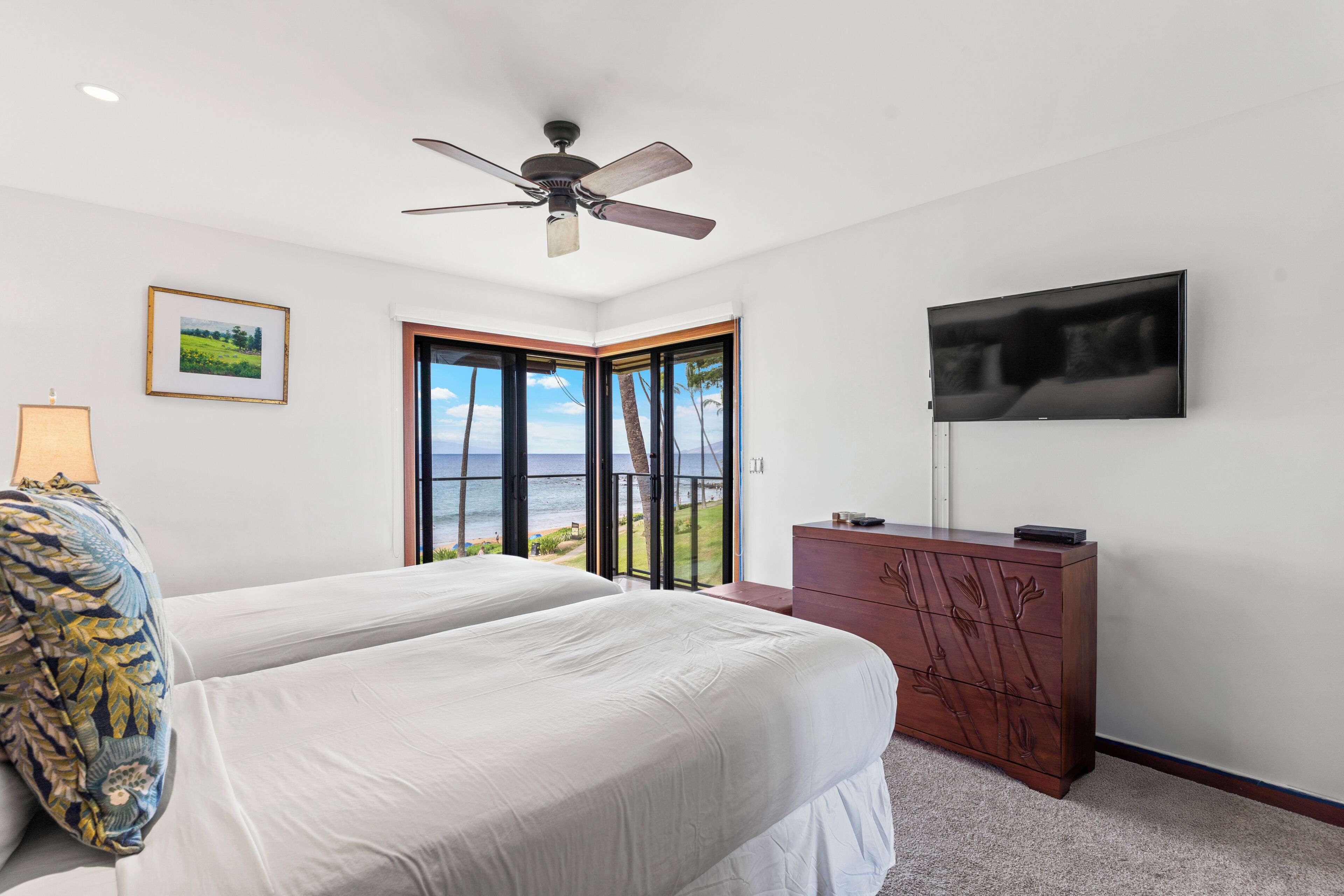 A bedroom with two beds, a ceiling fan, a TV, a dresser, and a large window with a view of the ocean.