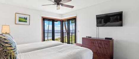 A bedroom with two beds, a ceiling fan, a TV, a dresser, and a large window with a view of the ocean.