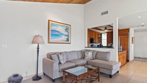 A living room with a sectional sofa, a glass coffee table, a lamp, and a painting of a beach scene. - Wailea Elua 1304 - Spectacular Oceanfront Condo with Panoramic Views (Wailea, Maui)
